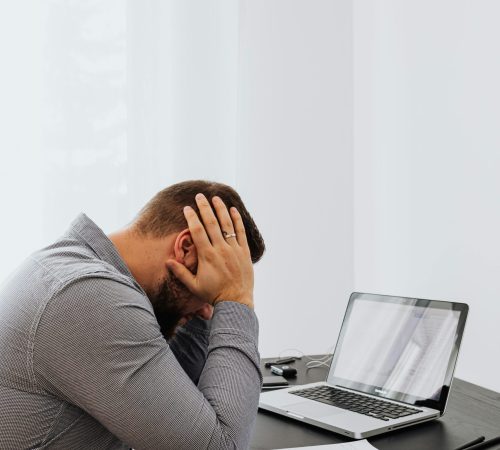 A frustrated man sits at a desk with a laptop and charts, showing work stress.